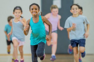 A group of elementary school children of various ethnicity are in PE class. They are indoors in the gym. They are playing a running game and smiling. A young girl of African descent is at the front.